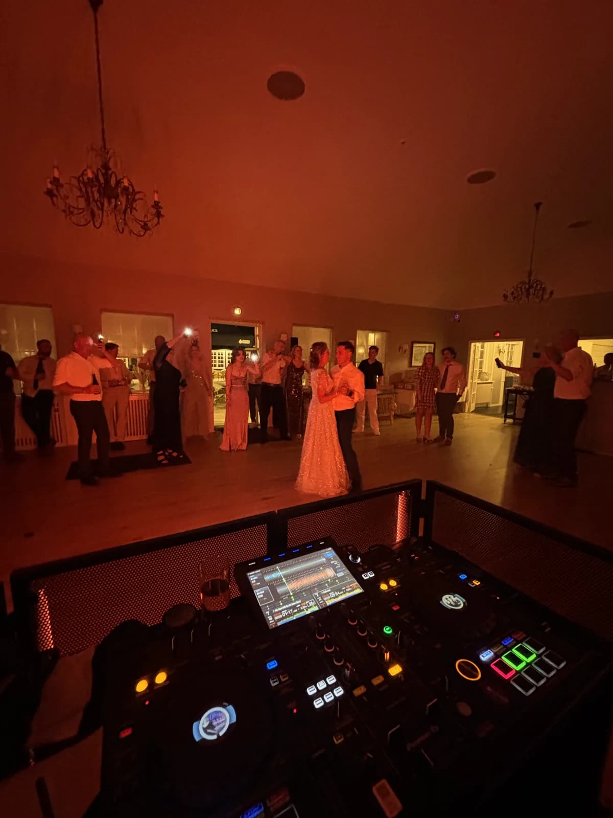 Brudevalsen set fra DJ-pulten: brudeparret danser alene midt på gulvet i varmt orange lys, mens gæsterne står i en ring langs væggene og filmer med telefonerne. To lysekroner hænger fra det hvælvede loft, og forrest i billedet ses mixeren med oplyste knapper.