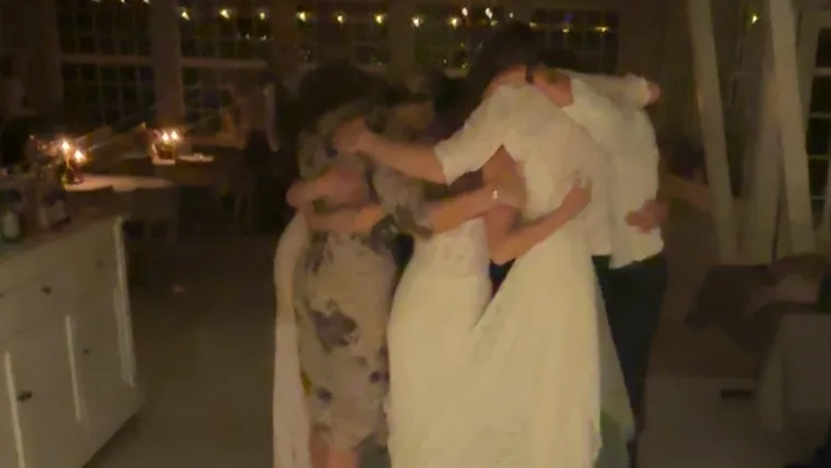 Fem gæster står i en tæt kreds midt på det lyse dansegulv med armene om hinanden sidst på aftenen. Bruden i hvid blondekjole står med ryggen til mellem en kvinde i blomstret kjole og en mand i hvid skjorte. Bag dem hænger en lyskæde i de høje sprossede vinduer, og til venstre står et hvidt barskab med levende lys.