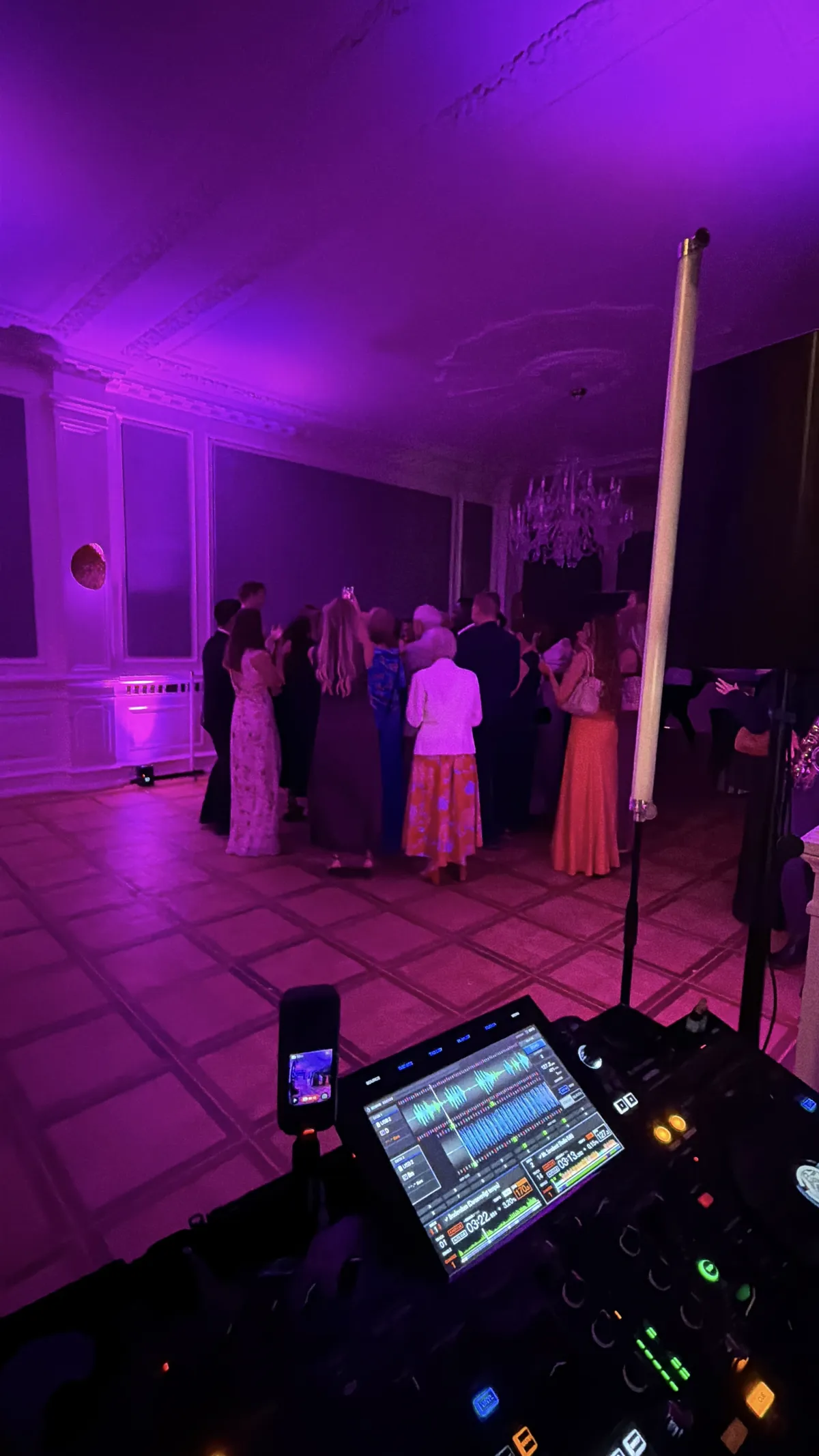 Set hen over DJ-pulten står omkring femten bryllupsgæster i lange kjoler og jakkesæt samlet på det lyse dansegulv i Hestkøbgaards sal. Loftet har stuk og en krystallysekrone, og hele rummet står i lilla og pink lys fra uplights langs væggene. Nederst i billedet lyser pultens skærm med et bølgeform-display.