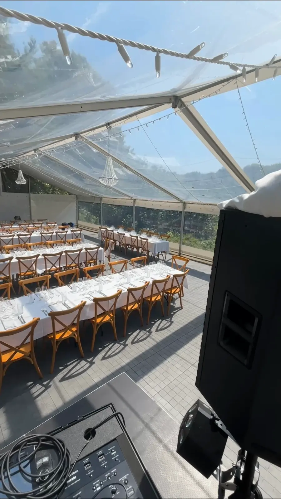 Teltet på Kongekilden set bag DJ-pulten om eftermiddagen, før gæsterne kommer. Gennemsigtigt tag med lyskæder mod en blå sommerhimmel, lange borde med hvide duge og træstole i rækker, og en sort højttaler på stativ ved pulten.