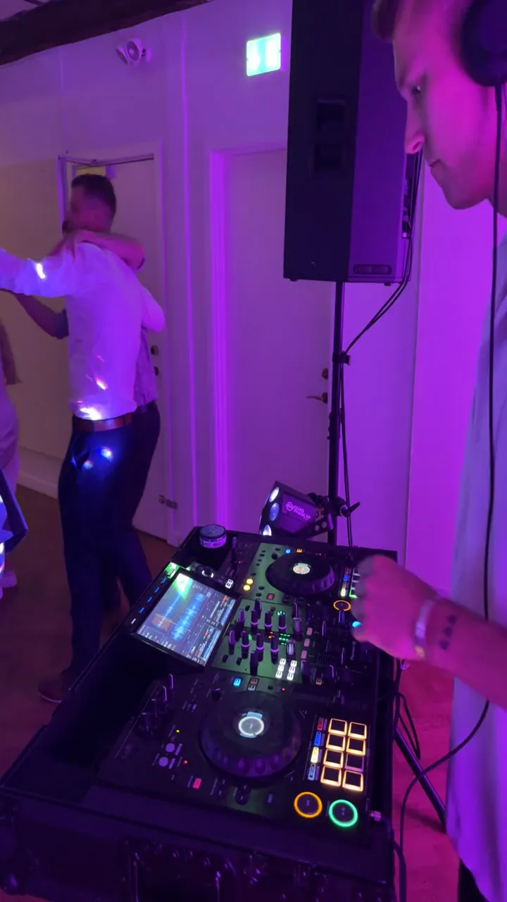 Set ved siden af DJ-pulten i et lyst lokale badet i lilla lys. Til højre står DJ'en med høretelefoner og hånden på pulten, i midten en højttaler på stativ, og til venstre danser to mænd i hvide skjorter tæt sammen med armene om hinanden.