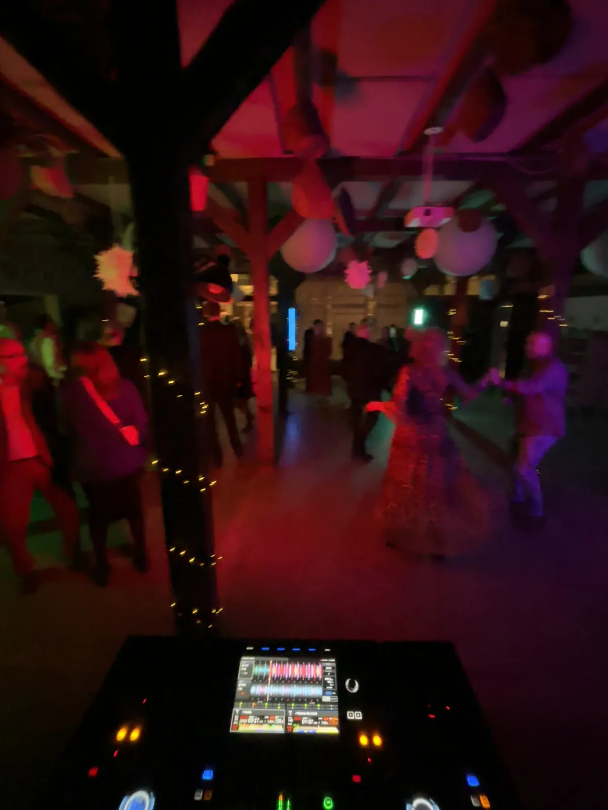 Set hen over DJ-pulten danser gæster i festtøj på det mørke gulv i en gammel trælade på BaneGaarden. Kraftige træsøjler bærer loftet, en lyskæde er viklet om søjlen i midten, og der hænger store hvide papirkugler mellem bjælkerne. Rummet er lyst op i rødt og lilla.