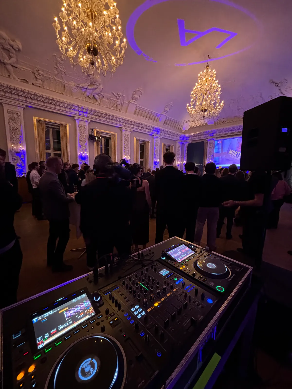 Set fra DJ-pulten på Moltkes Palæ: en Pioneer-pult forrest og bag den en sal fuld af gæster i mørkt tøj under to store krystallysekroner og et hvidt stukloft med figurer. Partiets logo er projiceret i lilla i loftet, væggene er lyst blå op, og i baggrunden viser en storskærm valgaftenens tv.