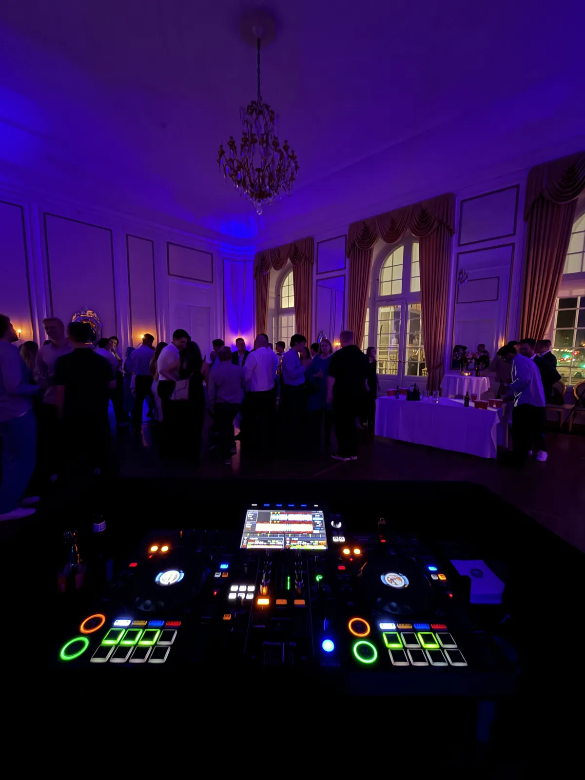 Festsalen på Odd Fellow set fra DJ-pulten i lilla lys: gæsterne står og snakker under en stor lysekrone, ved de høje vinduer står et dækket bord, og forrest lyser pultens knapper i grønt og orange.