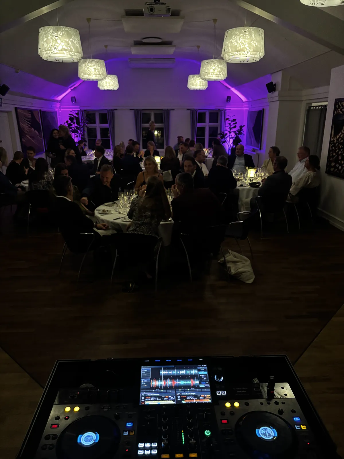 Set hen over DJ-pulten står dansegulvet af mørkt træ tomt, mens 70 gæster i festtøj sidder til middag ved runde borde med hvide duge og vinglas. Salen har store hvide lampeskærme i loftet, og væggene er lyst op i lilla. Nederst i billedet lyser pultens skærm.