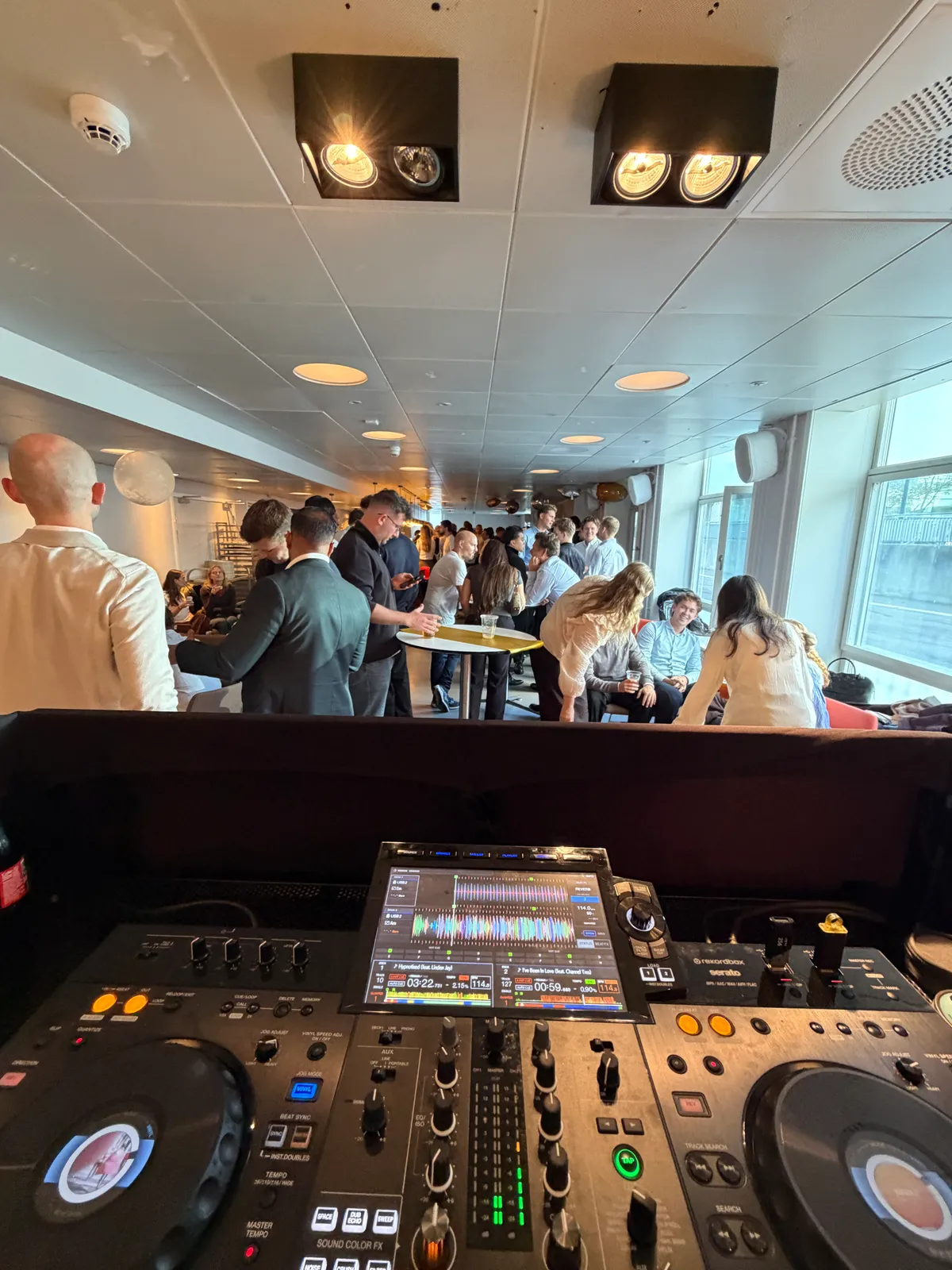 Set hen over DJ-pulten ud i fredagsbaren hos PwC: unge kolleger i skjorter og jakkesæt står og snakker ved ståborde og sidder i sofaer i et lyst lokale med store vinduer, spots i loftet og hvide højttalere på væggen.