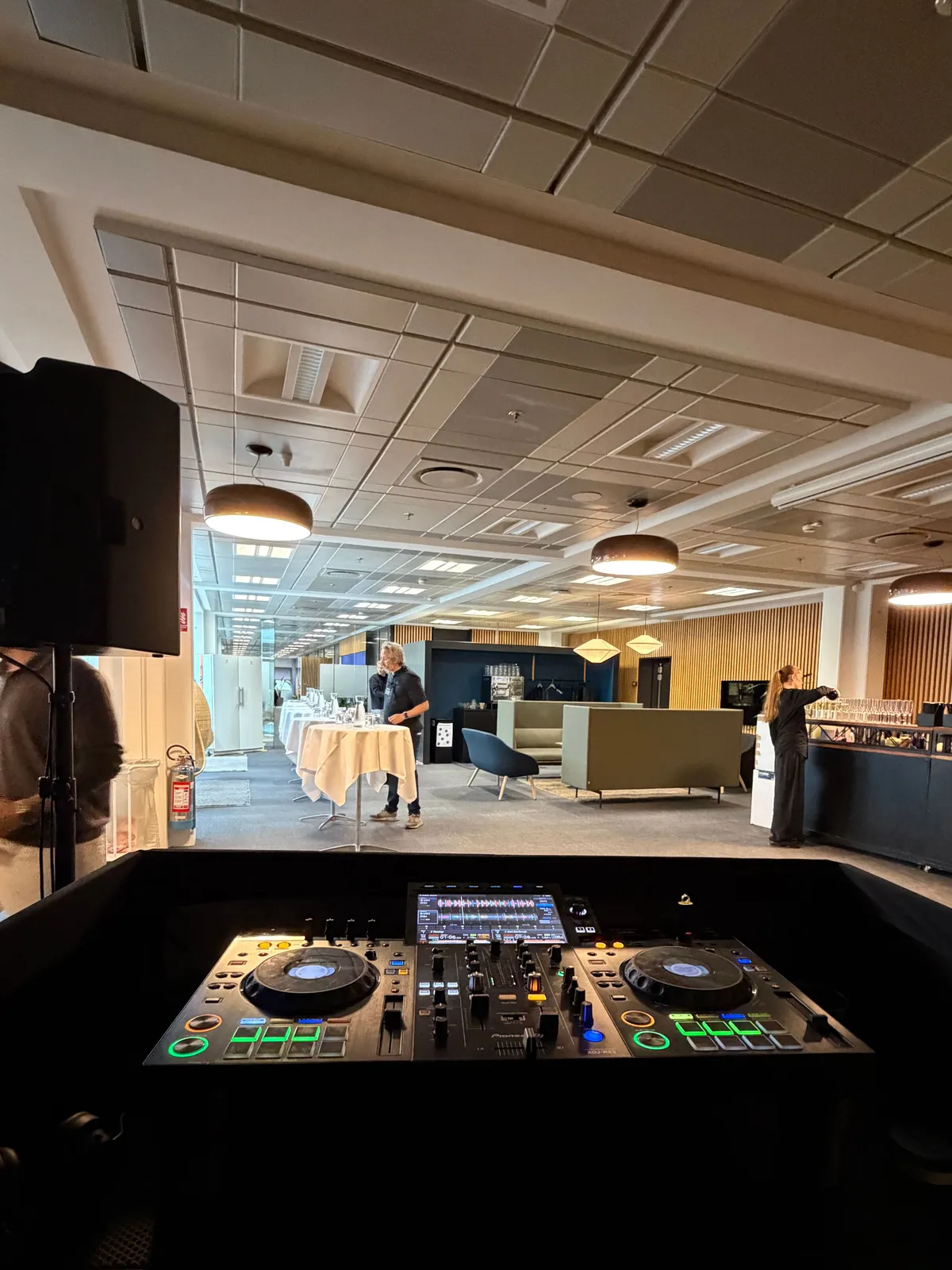 Set fra DJ-pulten, en Pioneer XDJ-RX3, ud over lokalet lige før s360s jubilæum: en højttaler på stativ, hvide ståborde med duge, grønne sofaer og en lang bar med glas, hvor personalet gør klar, i et lyst kontorlokale med lamper i loftet og træpaneler på væggen.