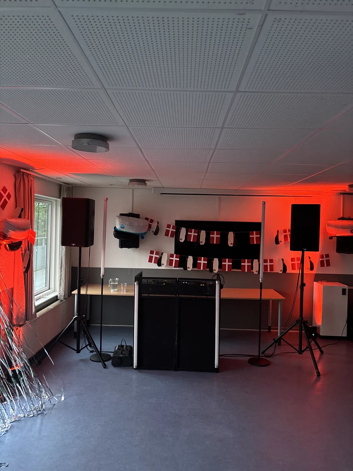 Det lange festlokale før gæsterne kommer: guirlander med Dannebrogsflag og studenterhuer hænger på tværs under et hvidt akustikloft, DJ-pulten står klar med to højttalere og lysstave, og væggene er badet i rødt lys.