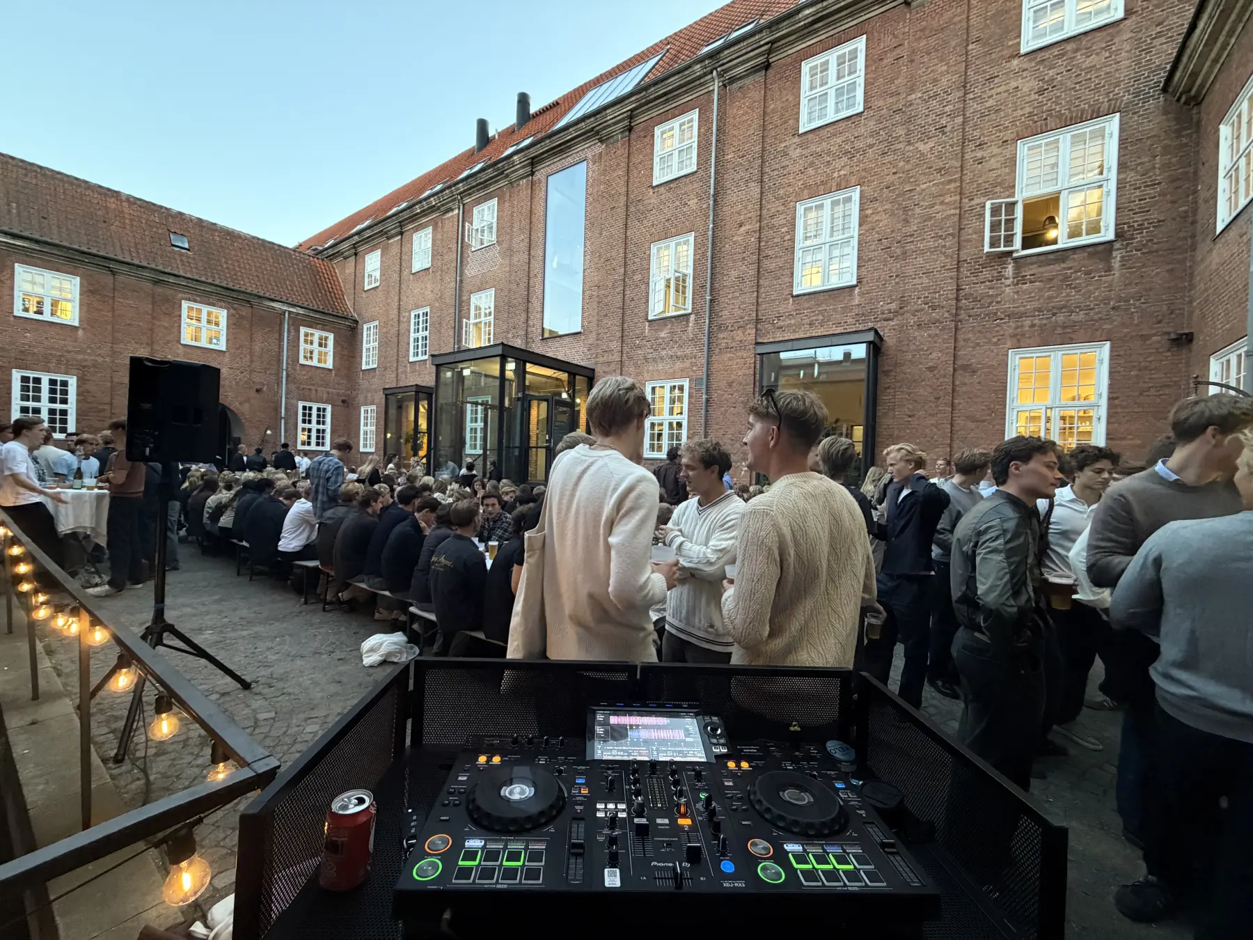 Set fra DJ-pulten ud over en brostensbelagt gård i København: studerende står med øl i hånden foran lange spiseborde, og lyskæder hænger langs den røde murstensbygning bag dem.