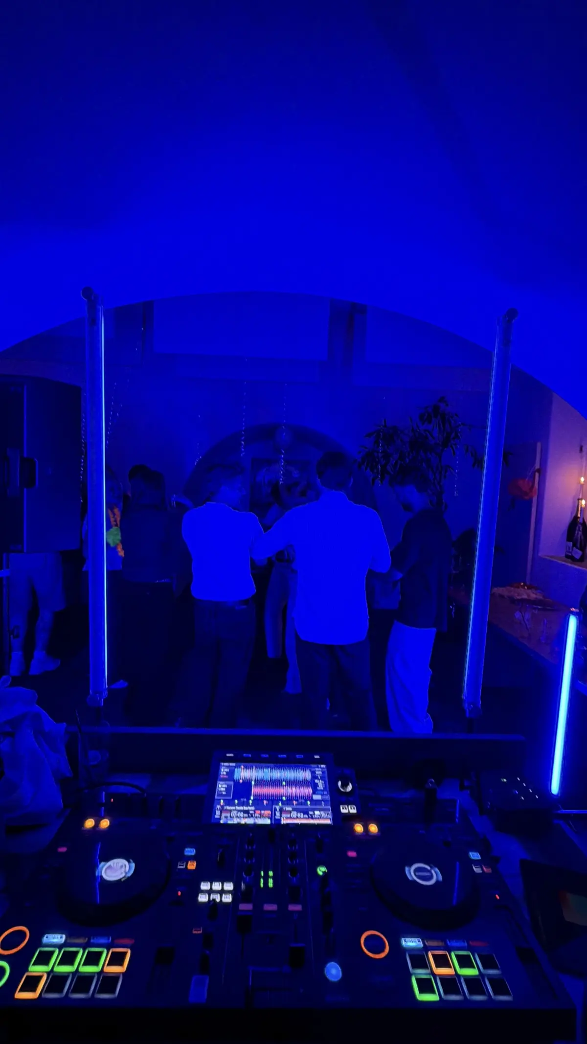 Set fra DJ-pulten ud mod dansegulvet i blåt lys: en lille gruppe gæster står med ryggen til mellem to lodrette lysstave, under et hvælvet loft.