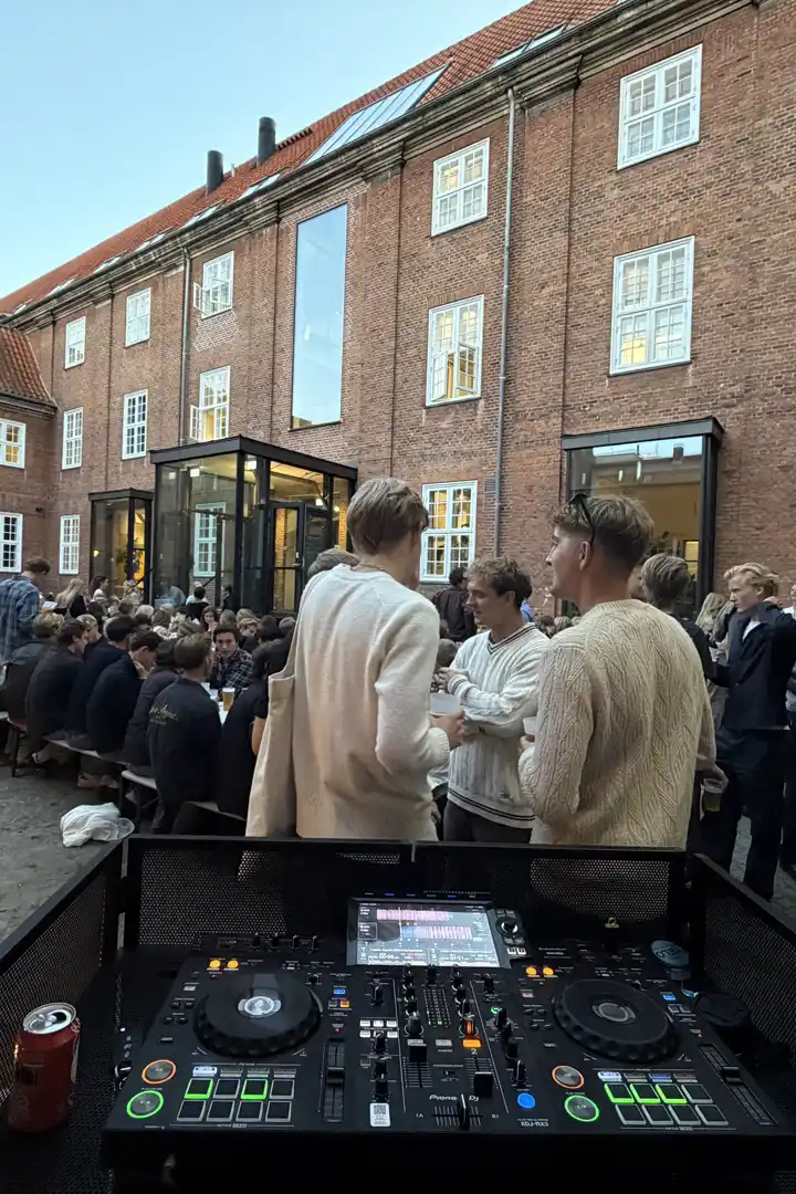 Gæster der står og snakker i en brolagt gård foran en murstensbygning, set hen over DJ-pulten i aftenlys